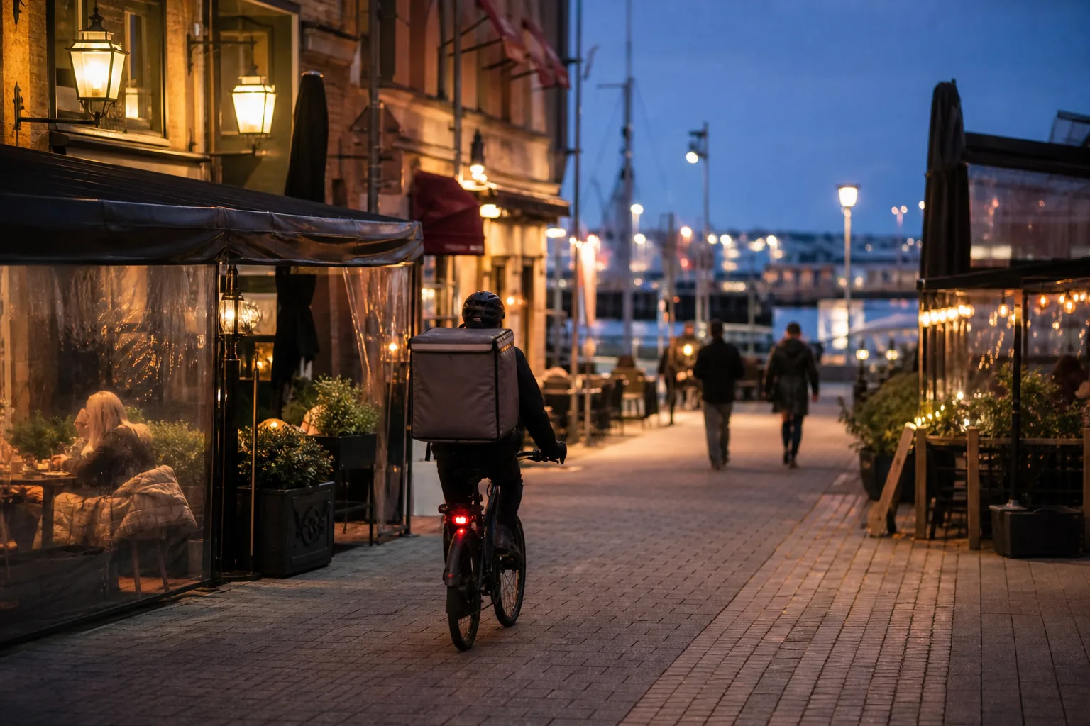 Illustrativ kvällsbild av en svensk hamnstad med en cykelbud på väg mellan restauranger